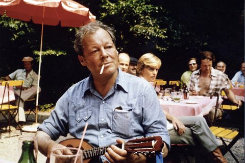Willy Brandt sitzt und spielt Mandoline mit Zigarette im Mund, im Hintergrund ein Sonnenschirm und Menschen an Tischen