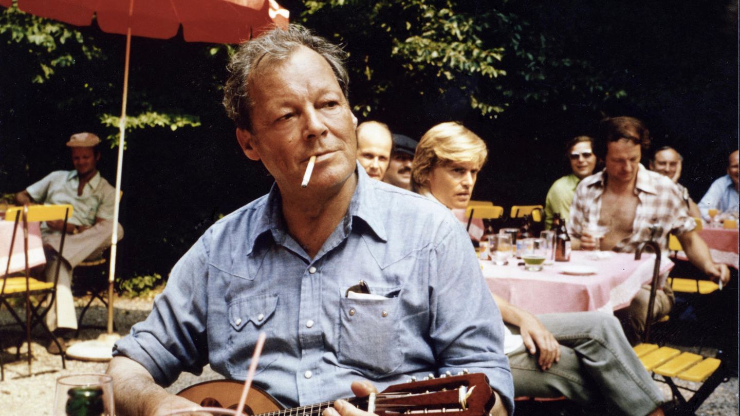 Willy Brandt sitzt und spielt Mandoline mit Zigarette im Mund, im Hintergrund ein Sonnenschirm und Menschen an Tischen