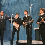Die Beatles mit Instrumenten auf einer Bühne, neben ihnen Ed Sullivan