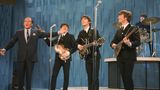Die Beatles mit Instrumenten auf einer Bühne, neben ihnen Ed Sullivan