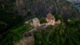 Luftaufnahme von einem Schloss auf einem bewaldeten Berg, umgeben von weiteren Bergen