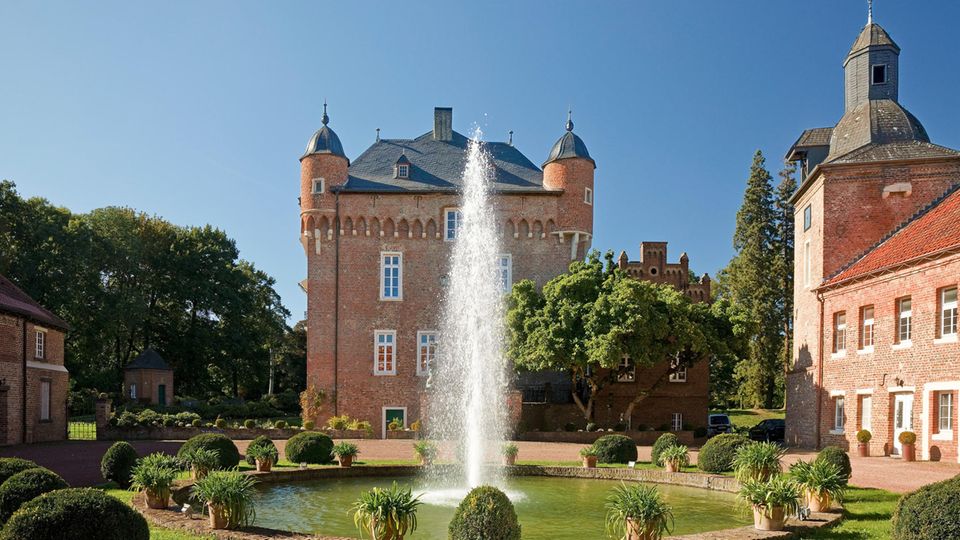 Rötliche Gebäude um einen Teich mit Wasserfontaine, drumherum Sträucher und Büsche Rötliche Gebäude um einen Teich mit Wasserfontaine, drumherum Sträucher und Büsche