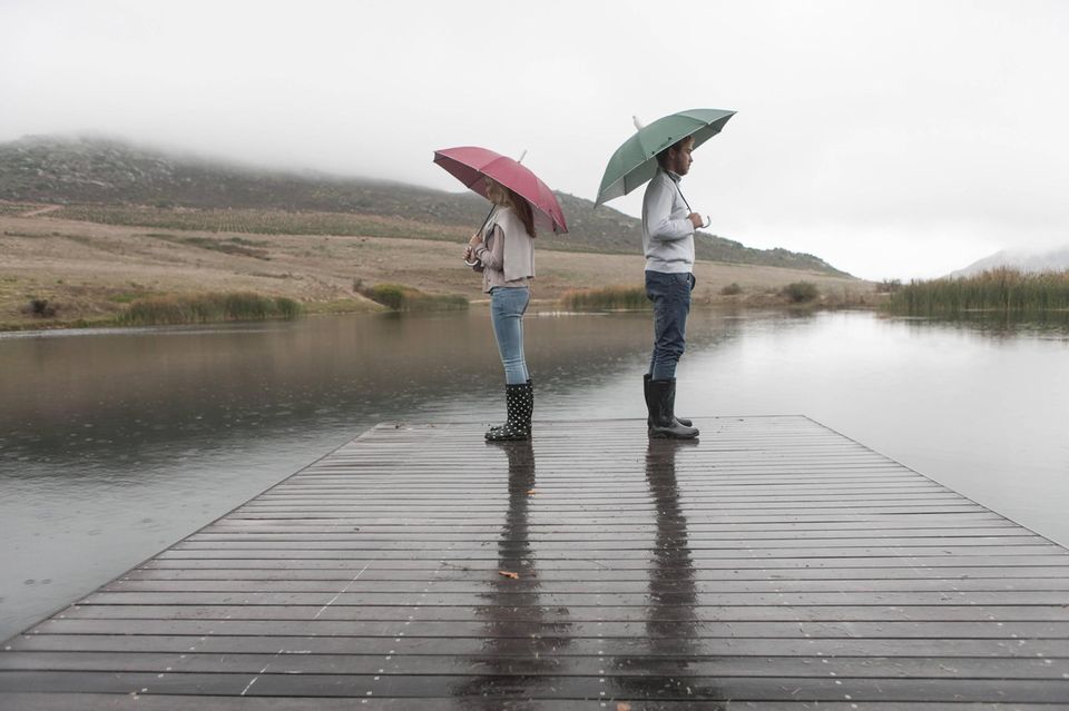 Ein Mann und eine Frau stehen Rücken an Rücken auf einem Steg mit Regenschirmen