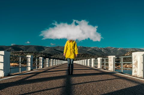 Frau mit gelben Regenmantel und einer Wolke vor dem Kopf steht auf einer Brücke