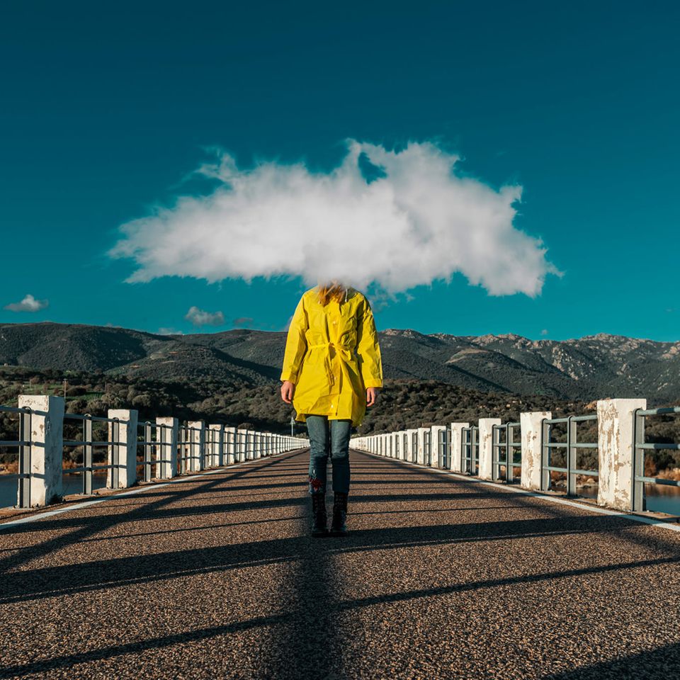 Frau mit gelben Regenmantel und einer Wolke vor dem Kopf steht auf einer Brücke