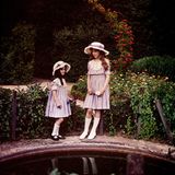 Zwei junge Mädchen in Kleidern lehnen an blumigen Büschen vor einem Teich