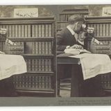 Mann schaut vor einem Bücherregal durch ein fernglasartiges Objekt, auf dem Schoß eine Weltkarte