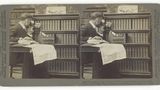 Mann schaut vor einem Bücherregal durch ein fernglasartiges Objekt, auf dem Schoß eine Weltkarte