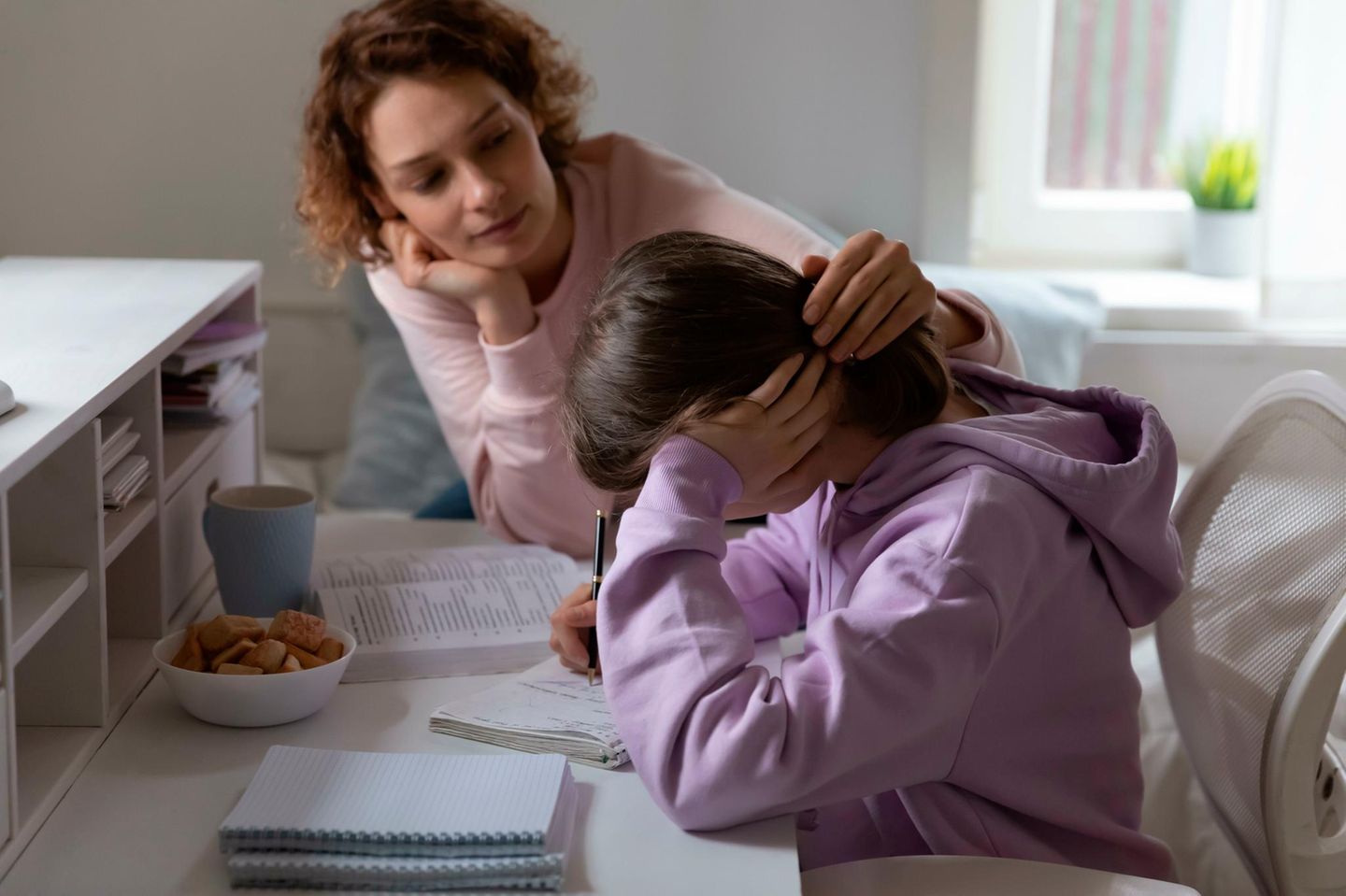 Mutter streichelt ihre gestresste Teenagertochter, die nach vorn gebeugt, am Schreibtisch sitzt und in ein Schulheft schreibt