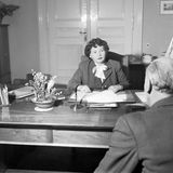 Schwarz-weiss Foto einer älteren Frau, die in Kostüm und Bluse an ihrem Schreibtisch sitzt im Gespräch mit einem Mann