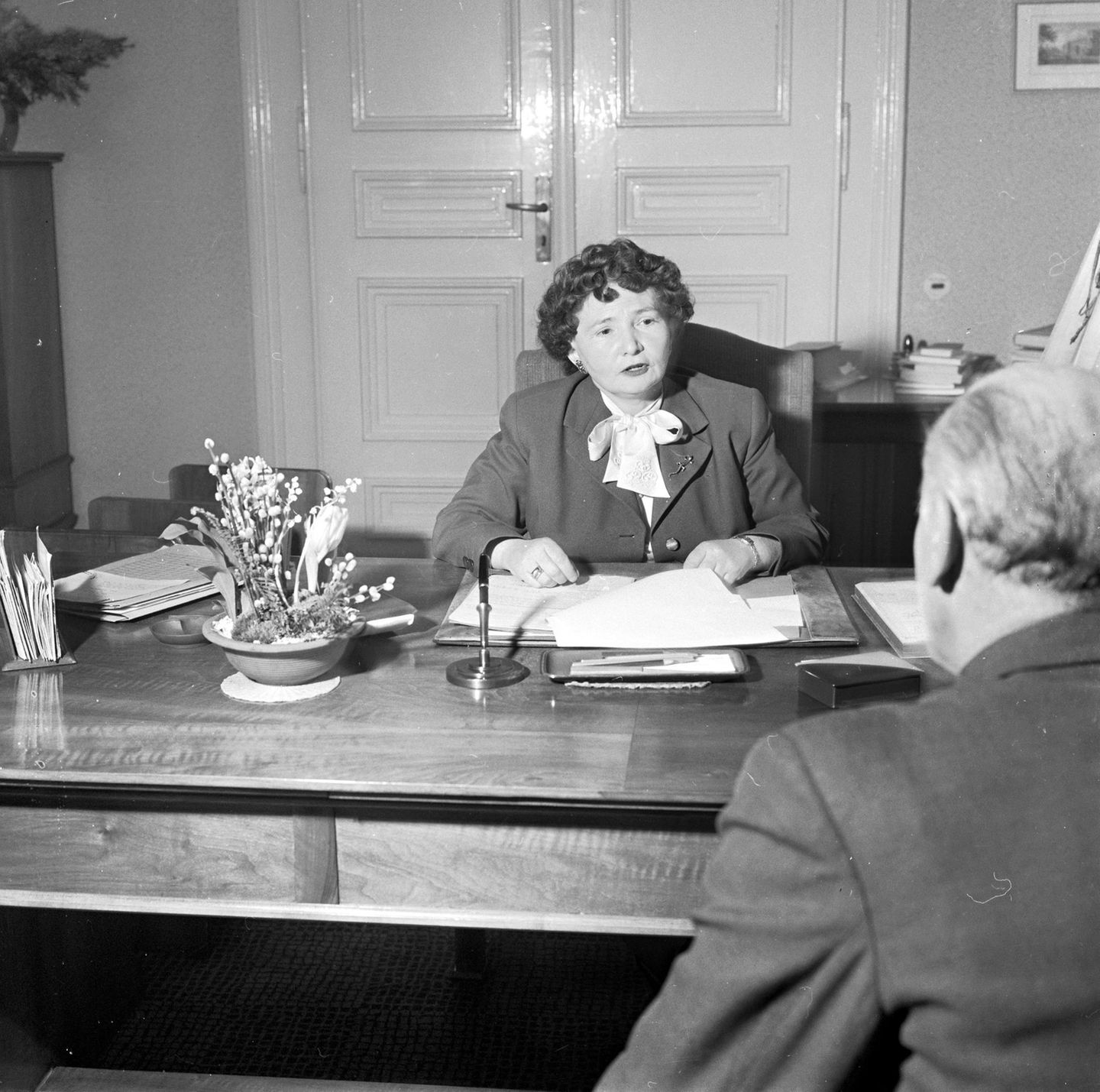 Schwarz-weiss Foto einer älteren Frau, die in Kostüm und Bluse an ihrem Schreibtisch sitzt im Gespräch mit einem Mann