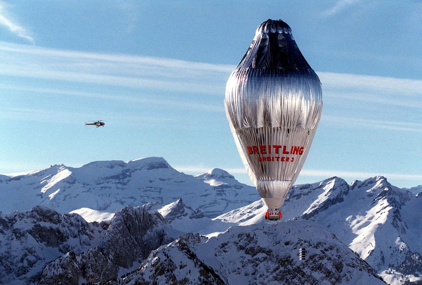 Ein silbern glänzender, länglicher Ballon mit einer roten Gondel überquerte schneebedeckte Alpengipfel