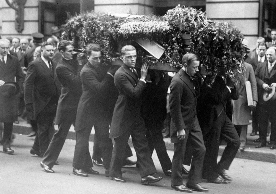 Acht Männer in dunklen Anzügen tragen Harry Houdinis blumenbedeckten Sarg durch eine Straße in New York