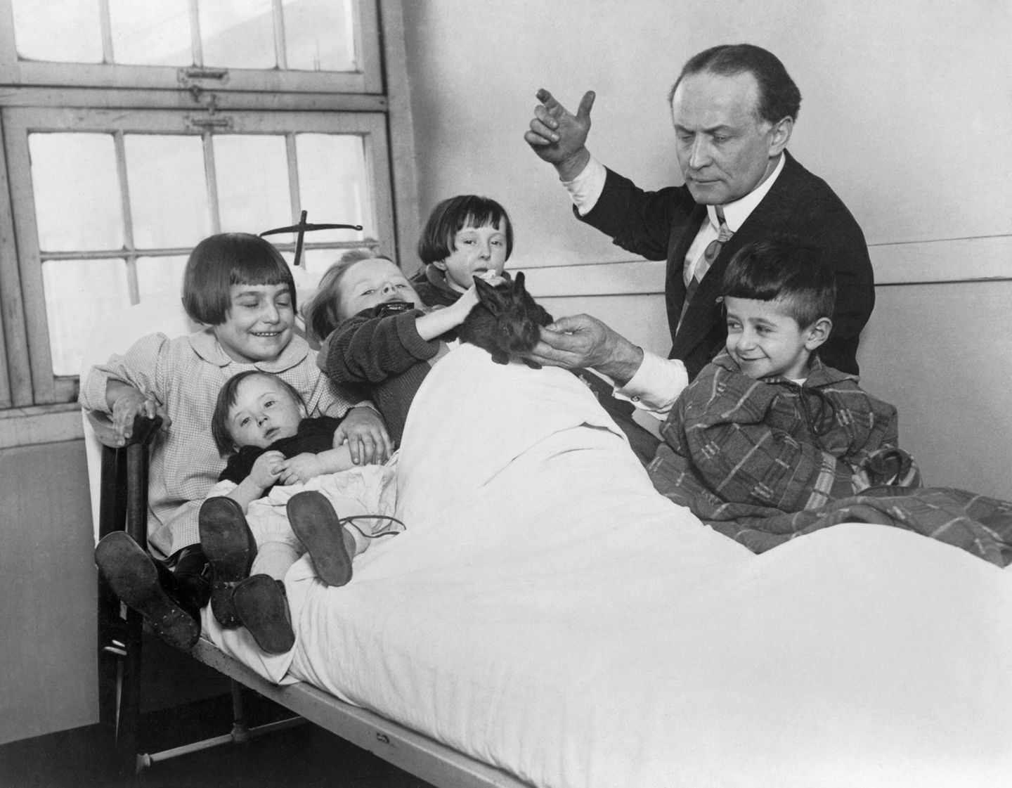 Houdini steht im Anzug an einem eisernen Krankenhausbett mit fünf Kindern und unterhält sie mit einem Kaninchen