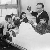 Houdini steht im Anzug an einem eisernen Krankenhausbett mit fünf Kindern und unterhält sie mit einem Kaninchen