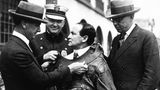 Drei schmunzelnde Polizisten fesseln Harry Houdini mit einer Zwangsjacke