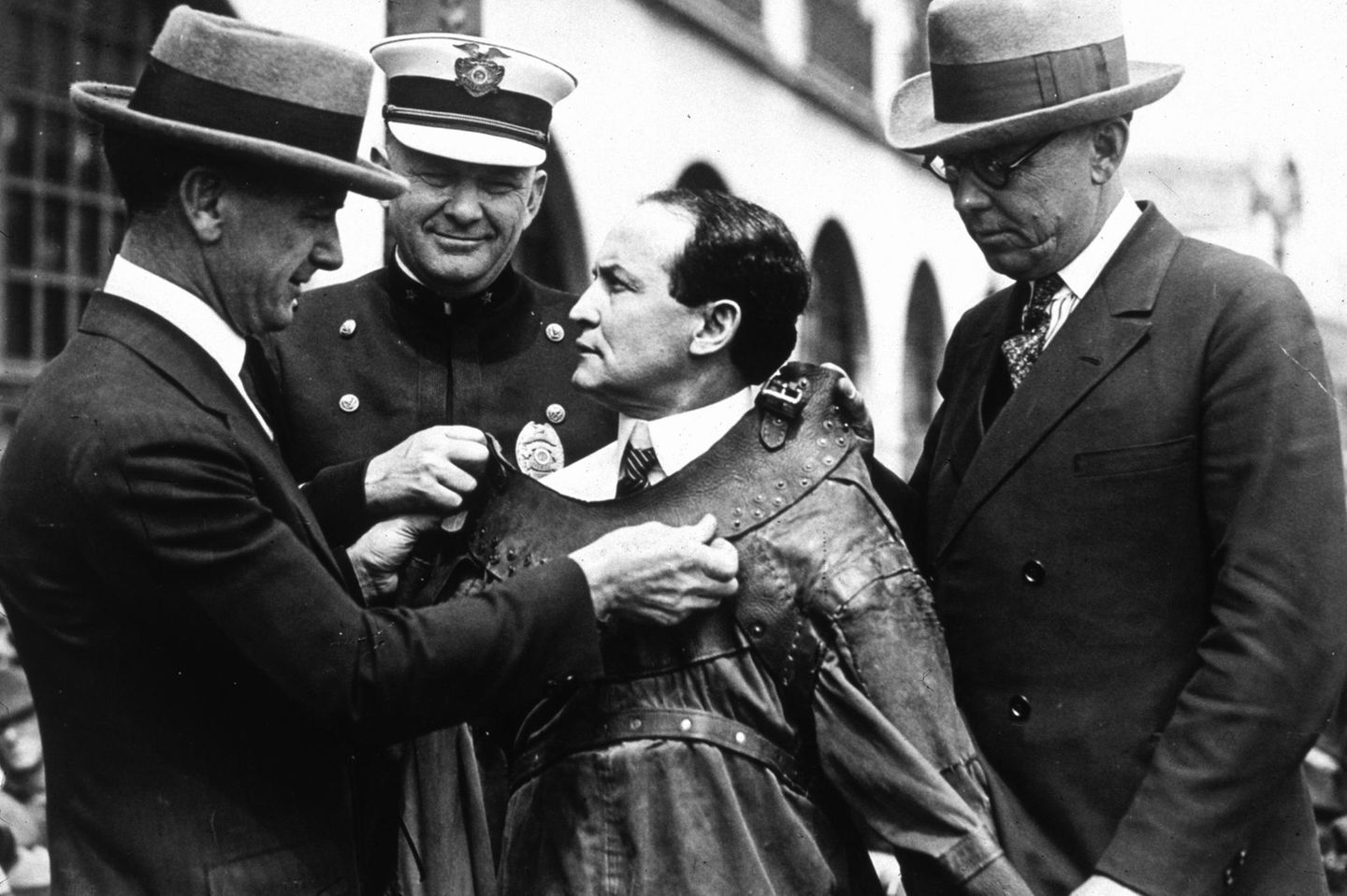 Drei schmunzelnde Polizisten fesseln Harry Houdini mit einer Zwangsjacke