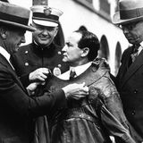 Drei schmunzelnde Polizisten fesseln Harry Houdini mit einer Zwangsjacke