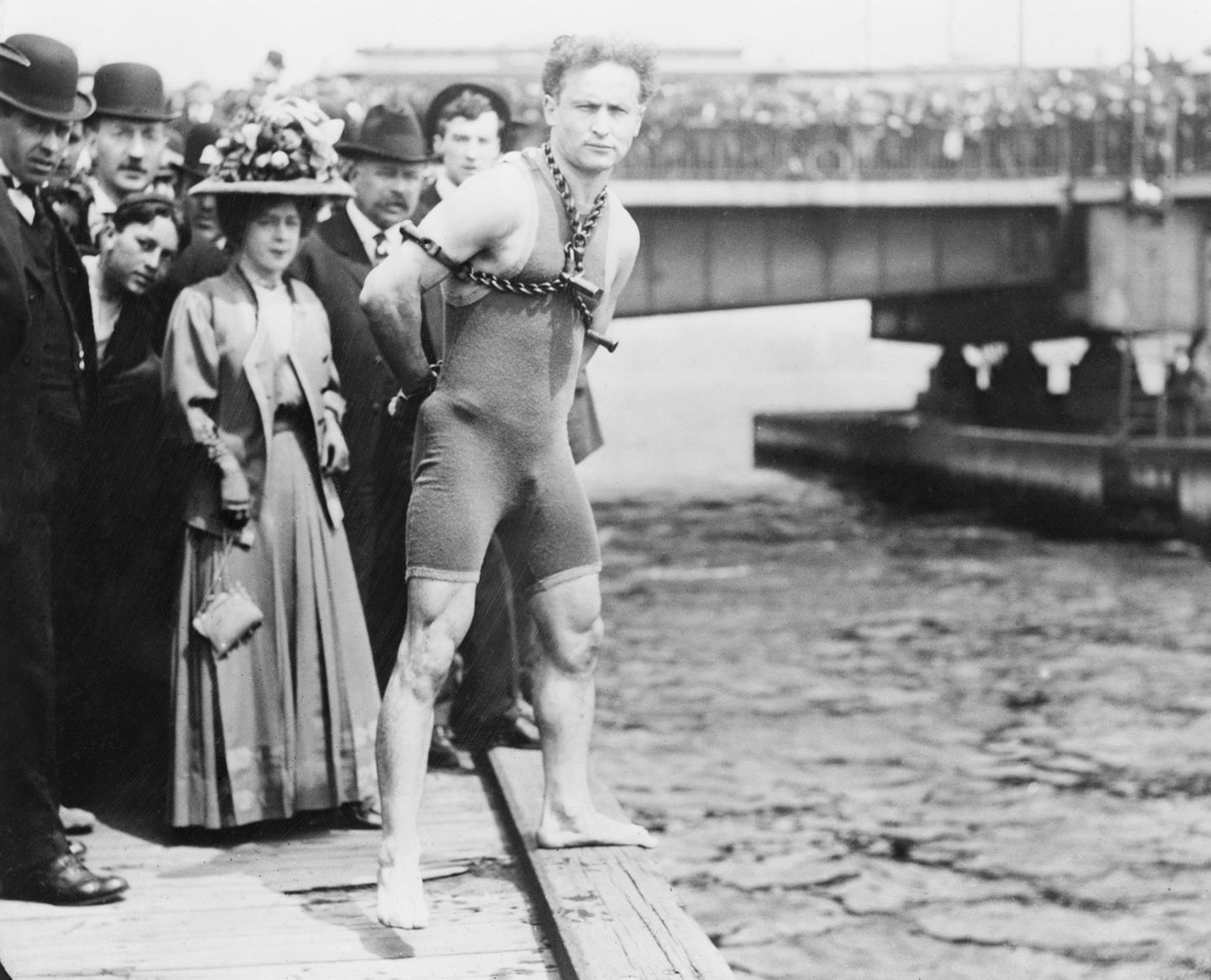 Harry Houdini posiert gefesselt und im Badeanzug am Ufer des Charles River, Boston. Im Hintergrund eine Menschenmenge.