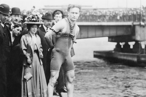 Harry Houdini posiert gefesselt und im Badeanzug am Ufer des Charles River, Boston. Im Hintergrund eine Menschenmenge.