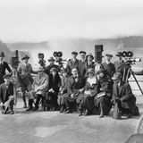 15 Männer und Frauen in Mänteln und Hüten posieren in zwei Reihen vor den Niagara-Fällen, zwischen ihnen Filmkameras.