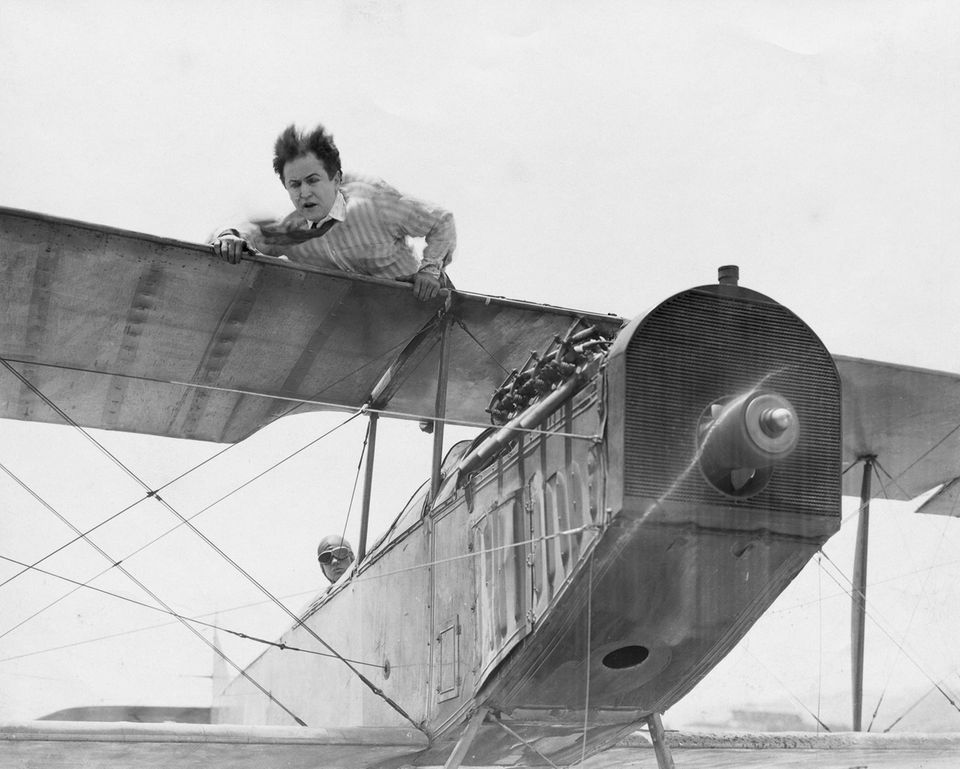 Houdini hält sich hoch in der Luft mit wehenden Haaren auf der Tragfläche eines alten, kleinen Flugzeugs fest.