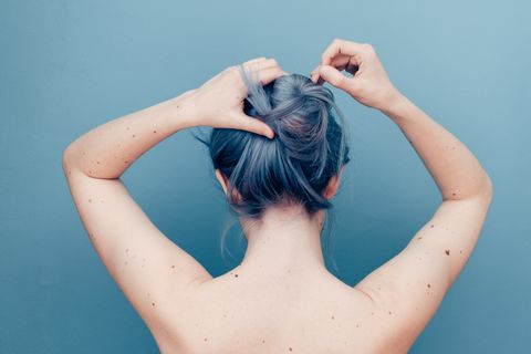 Rückenporträt einer jungen entblößten Frau mit blau gefärbten Haaren, die sich mit erhobenen Armen eine Frisur macht.