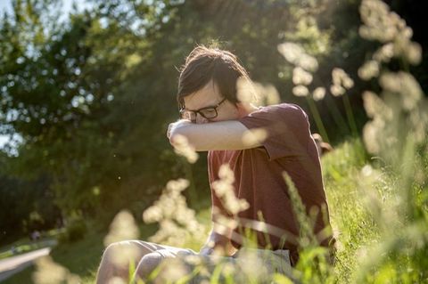 Ein Mann sitzt auf einer Blumenwiese und niest in die Armbeuge