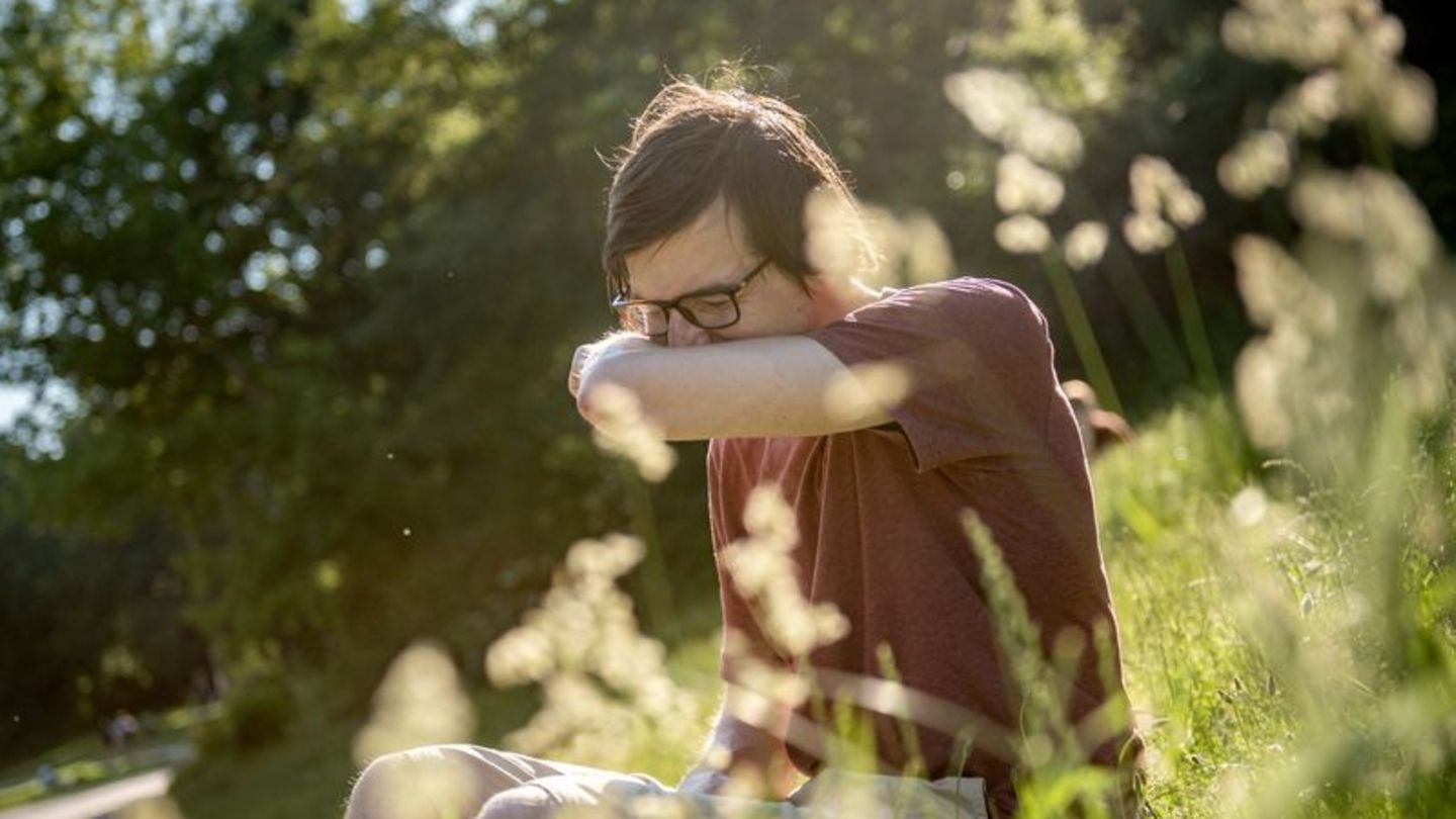 Ein Mann sitzt auf einer Blumenwiese und niest in die Armbeuge