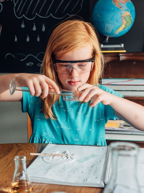 Ein Mädchen hantiert vor einer Schultafel mit Reagenzglas, umgeben von wissenschaftlich anmutenden Utensilien