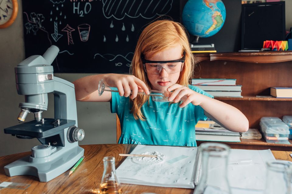 Ein Mädchen hantiert vor einer Schultafel mit Reagenzglas, umgeben von wissenschaftlich anmutenden Utensilien