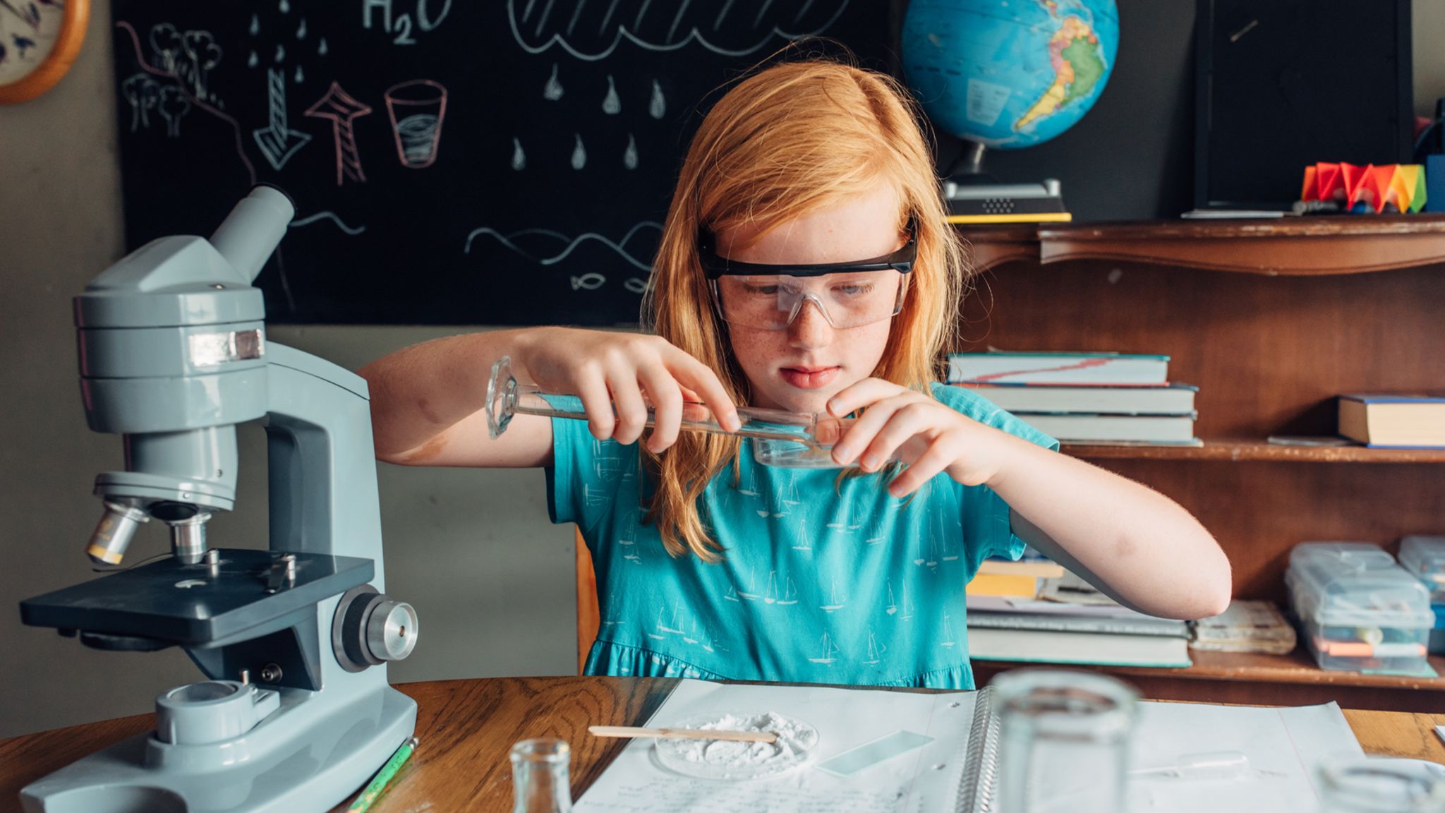 Ein Mädchen hantiert vor einer Schultafel mit Reagenzglas, umgeben von wissenschaftlich anmutenden Utensilien