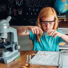 Ein Mädchen hantiert vor einer Schultafel mit Reagenzglas, umgeben von wissenschaftlich anmutenden Utensilien