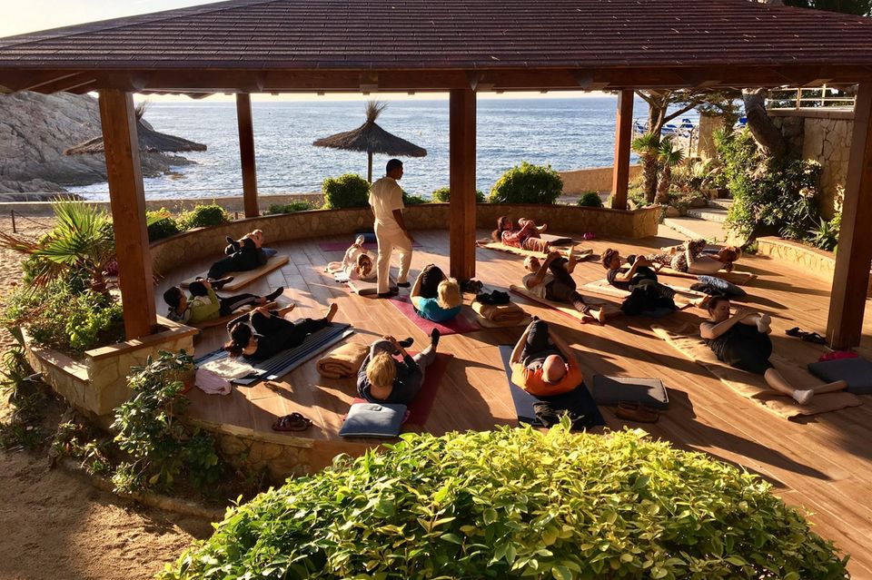 Menschen machen Yoga auf Yogamatten unter Sonnenschirmen auf Terrasse, dahinter das Meer 1.3333