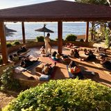 Menschen machen Yoga auf Yogamatten unter Sonnenschirmen auf Terrasse, dahinter das Meer