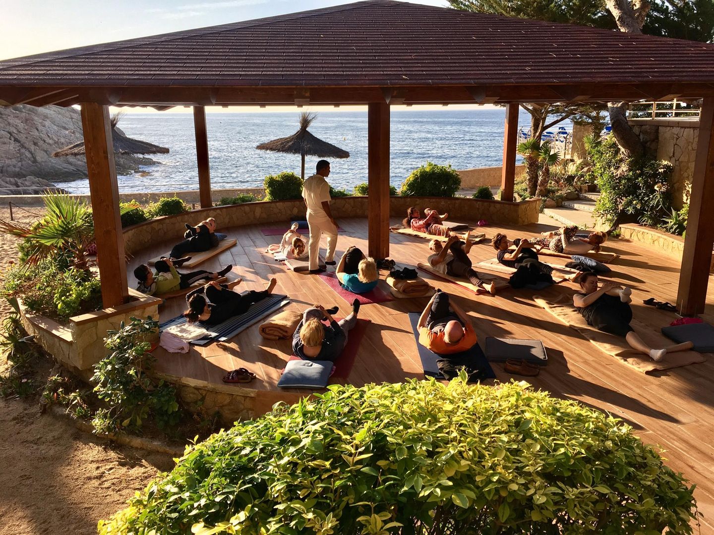 Menschen machen Yoga auf Yogamatten unter Sonnenschirmen auf Terrasse, dahinter das Meer