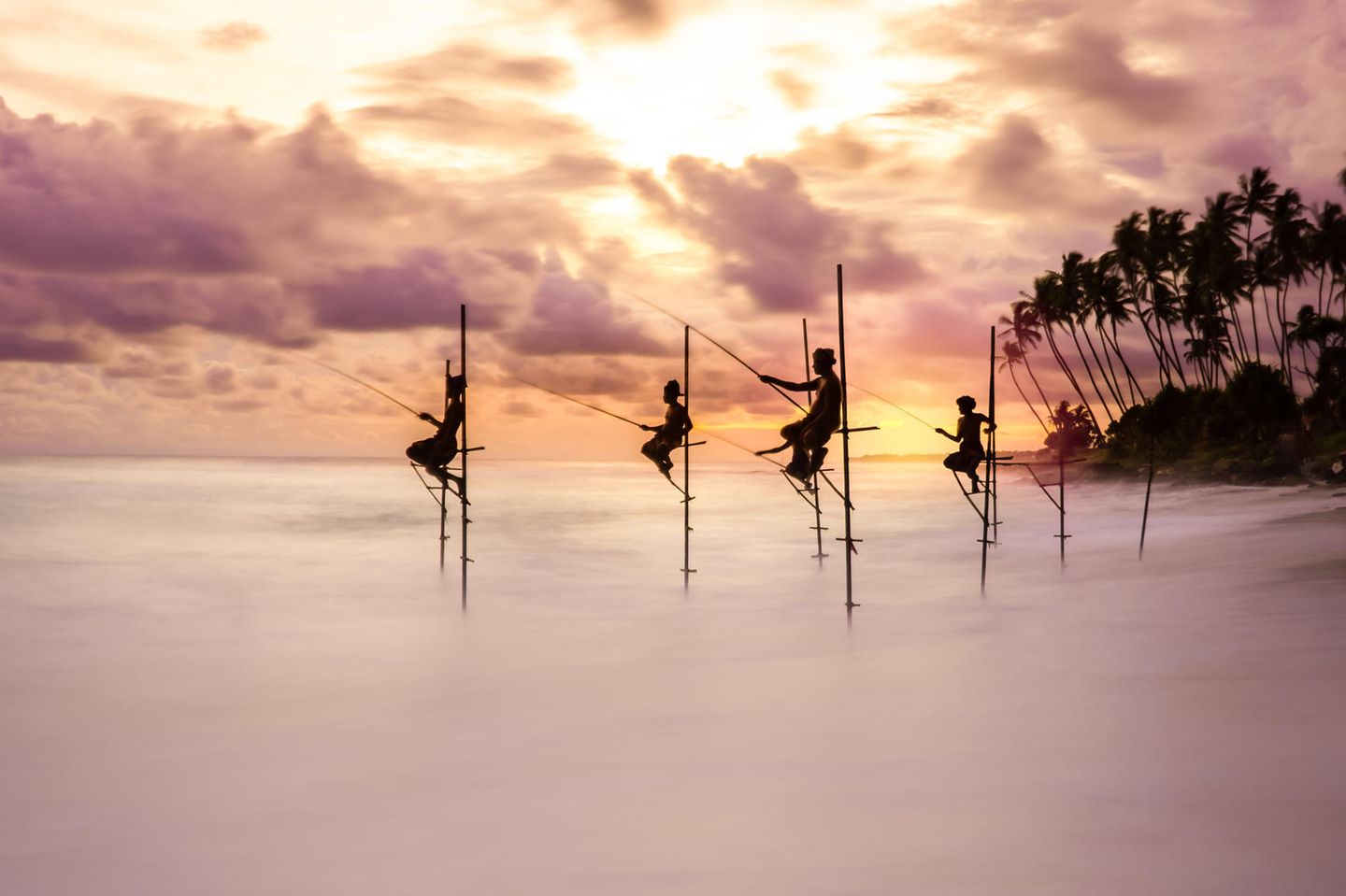 Traditionelle Stelzenfischer versuchen ihr Glück im Wechsel zwischen Ebbe und Flut bei Sonnenuntergang in Koggala, Sri Lanka. Das ausdrucksstarke Bild zeigt die Bewegung des Wassers im Kontrast zur Stille der Fischer.