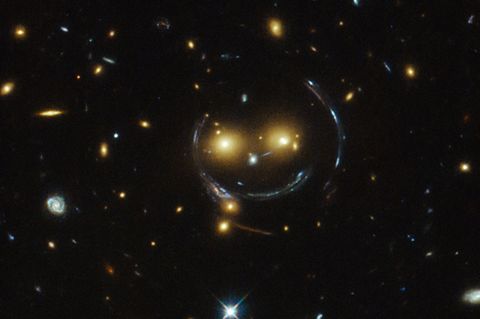 Der geisterhafte Smiley entsteht durch den Gravitationslinseneffekt: Die enorme Masse von Galaxieclustern krümmt die Raumzeit und damit auch die Lichtstrahlen der dahinterliegenden Galaxien. Diese werden im Bild zu einem kreisförmigen "Einsteinring" verzerrt