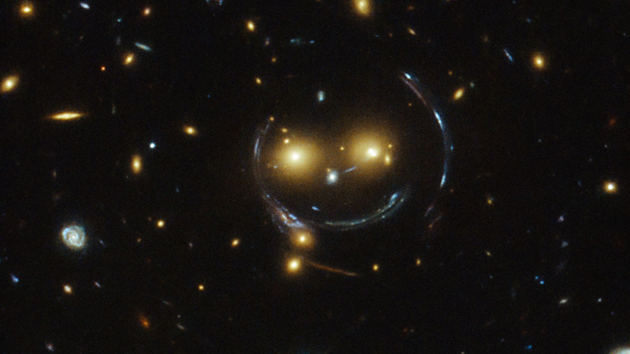 Der geisterhafte Smiley entsteht durch den Gravitationslinseneffekt: Die enorme Masse von Galaxieclustern krümmt die Raumzeit und damit auch die Lichtstrahlen der dahinterliegenden Galaxien. Diese werden im Bild zu einem kreisförmigen "Einsteinring" verzerrt