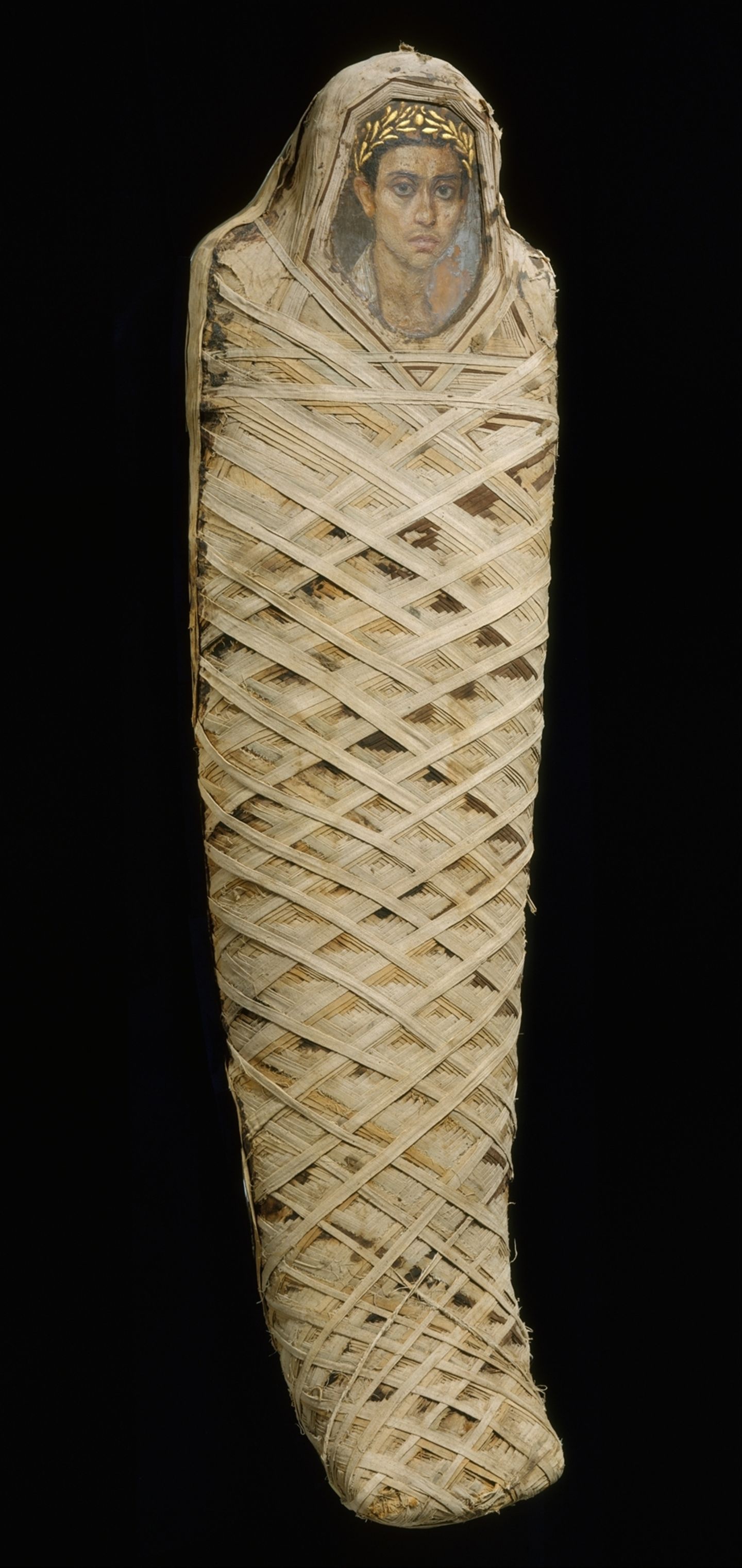 Eine mit Leinenbandage umwickelte Mumie, deren Gesicht von einem Porträtbild eines jungen Mannes bedeckt wird