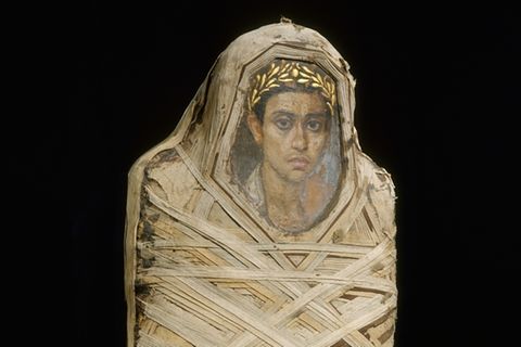 Eine mit Leinenbandage umwickelte Mumie, deren Gesicht von einem Porträtbild eines jungen Mannes bedeckt wird