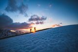 Zwei Leute wandern in der Abenddämmerung ein Gebirge hoch.