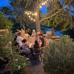 Familie sitzt unter altem Baum und Lichtergirlanden beim Abendessen.