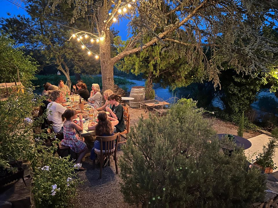 Familie sitzt unter altem Baum und Lichtergirlanden beim Abendessen.