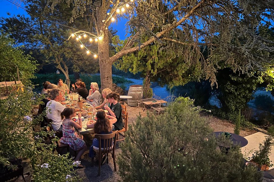 Familie sitzt unter altem Baum und Lichtergirlanden beim Abendessen.  1.3333