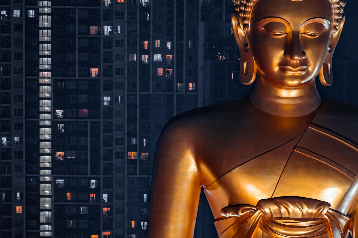 übergroße glänzendgoldene Buddhastatue, in der Nacht angeleuchtet. Dahinter Hochhaus mit beleuchteten Fenstern