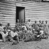 Eine Gruppe von Personen steht und sitzt vor einem Holzhaus