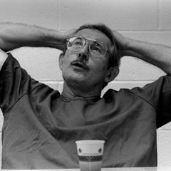 Ein Mann mit Schnurrbart und Brille sitzt im T-Shirt vor einer weißen Wand, die Arme hinter dem Kopf verschränkt