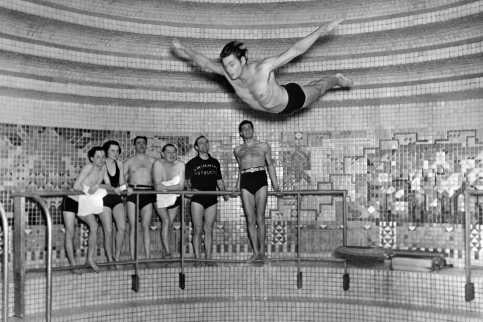 Johnny Weissmüller springt, beobachtet von einigen Personen, einen vollendeten Kopfsprung in ein Schwimmbecken