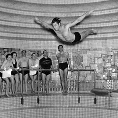 Johnny Weissmüller springt, beobachtet von einigen Personen, einen vollendeten Kopfsprung in ein Schwimmbecken